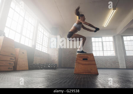 Vista laterale immagine di montare la giovane donna facendo un salto in scatola di esercizio. Donna muscolare facendo una scatola squat presso la croce montare palestra Foto Stock