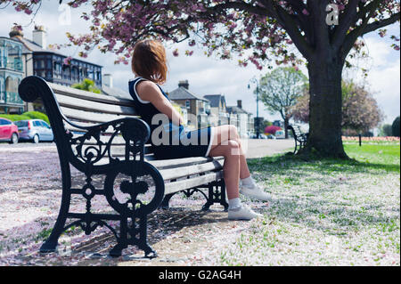 Un elegante giovane donna è seduta su una panchina nel parco con fiori di ciliegio sul terreno Foto Stock