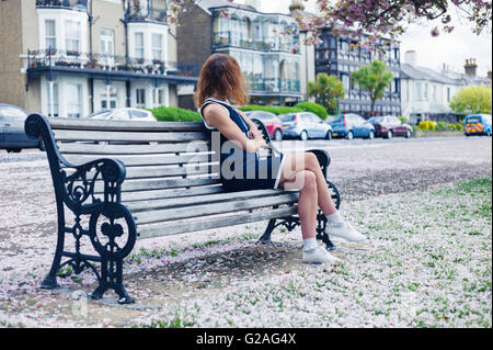 Un elegante giovane donna è seduta su una panchina nel parco con fiori di ciliegio sul terreno Foto Stock