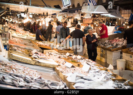 Mercato del pesce di Dimotiki Agora Atene Grecia | ATENE, Grecia - gli acquirenti si spostano tra casse di sardine e sgombri nella sala del pesce di Dimotiki Agora, sotto una bandiera greca e file di lampade da lavoro appese. Conosciuto anche come Varvakeios Agora, questo mercato del pesce coperto in via Athinas ha rifornito le famiglie ateniesi sin dall'apertura dell'edificio nel 1886 e rimane uno dei mercati di lavoro più trafficati della città. Foto Stock