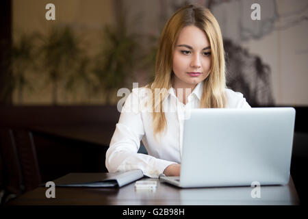 Giovane bella bionda imprenditrice caucasica lavorando su laptop, la digitazione con concentrati, di espressione cellulare e cartella nero Foto Stock