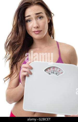 Close up studio shot di una giovane donna che guarda dietro una scala di peso su sfondo bianco con un'espressione preoccupata sul suo viso Foto Stock