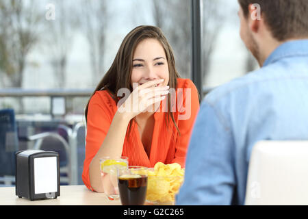 Donna che copre la bocca per nascondere un sorriso o alito cattivo durante una data in una caffetteria con una finestra in background Foto Stock