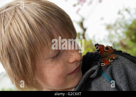 Ragazzo che guarda a farfalla pavone (Aglais io) seduto sulla sua spalla Foto Stock