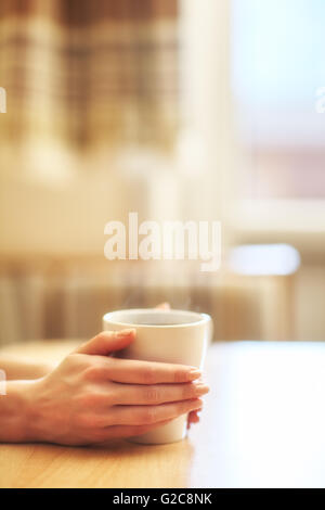 Le mani con la tazza di caldo il caffè al mattino sul tavolo con finestra in background Foto Stock