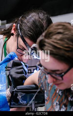 BELFAST, IRLANDA DEL NORD. 28 MAGGIO 2016 - 6th Conferenza del Tattoo dell'Irlanda del Nord. Un artista di tatuaggio disegna un tatuaggio intricato sul braccio di una cliente femminile con l'aiuto di una torcia da testa Foto Stock