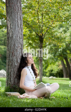 Giovane donna asiatica di sognare la posa contro un albero seduto in erba con un libro in mano Foto Stock