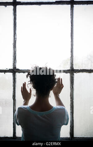 Vista posteriore di una giovane donna in piedi dalla finestra mani toccano il vetro Foto Stock