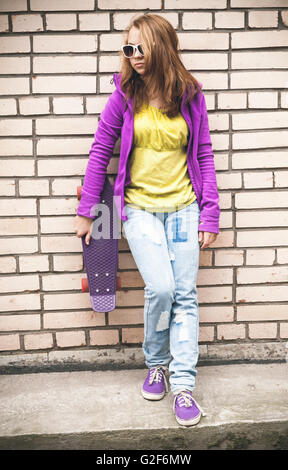 La bionda ragazza adolescente in occhiali da sole e abiti colorati con uno skateboard grigio vicino a un muro di mattoni Foto Stock