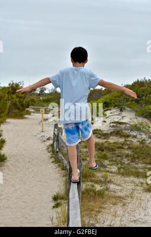 Un giovane ragazzo cerca di equilibrio come egli cammina attraverso la parte superiore di una recinzione di legno in spiaggia Foto Stock