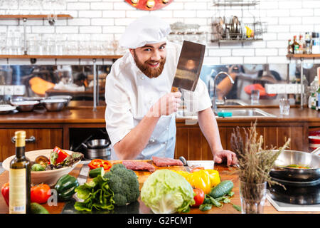 Sorridente attraente chef di cucina con coltello mannaia del taglio della carne e verdure in cucina Foto Stock