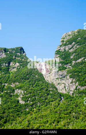 Ripide scogliere coperte di vegetazione con cascata in distanza che scorre dalla cima della montagna sotto il cielo blu chiaro. Foto Stock