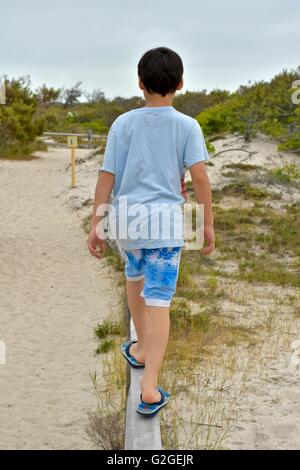 Un giovane ragazzo cerca di equilibrio come egli cammina attraverso la parte superiore di una recinzione di legno in spiaggia Foto Stock