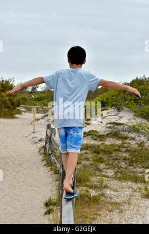 Un giovane ragazzo cerca di equilibrio come egli cammina attraverso la parte superiore di una recinzione di legno in spiaggia Foto Stock