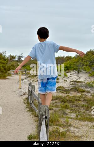 Un giovane ragazzo cerca di equilibrio come egli cammina attraverso la parte superiore di una recinzione di legno in spiaggia Foto Stock