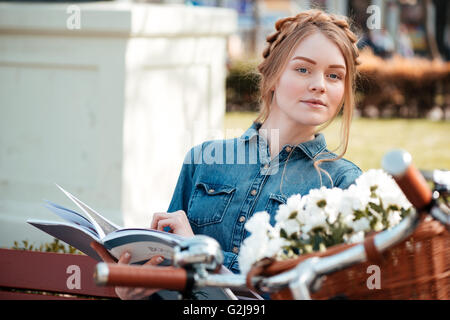 Bella ragazza bionda con bicicletta rivista di lettura sul banco di prova all'aperto Foto Stock