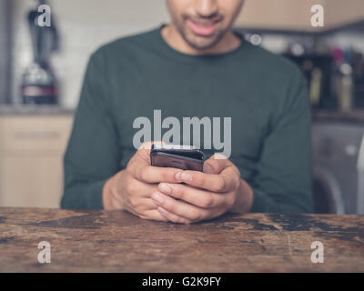 Un giovane uomo è seduta al tavolo in cucina e sta usando un telefono intelligente Foto Stock