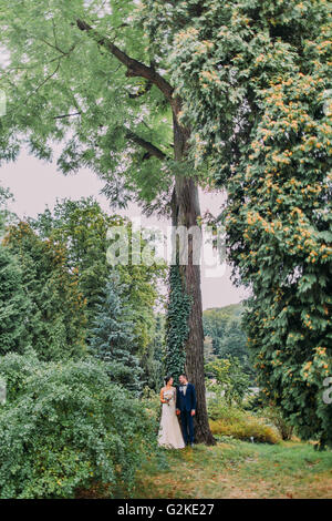 Appena sposata giovane sotto alta acacia-tree strisciato con ivy. Appena sposata con passeggiate nel parco nel giorno del loro matrimonio Foto Stock