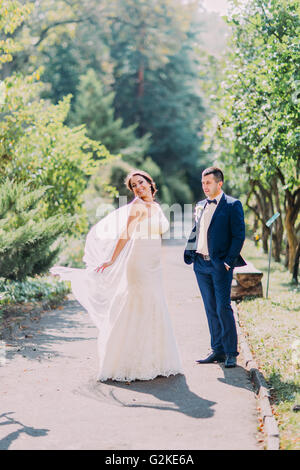 Sposi in posa soleggiato parco. Giocoso sposa che mostra il suo velo nuziale Foto Stock