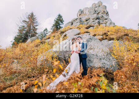 Bello lo sposo in elegante tuta blu abbracciando vestito di bianco sposa azienda bouquet di rose sul idilliaco paesaggio outdoor con grandi rocce come backround. Basso angolo di visione Foto Stock