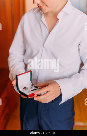 Attraente Giovane Uomo in camicia bianca tenendo stylish RED scatola nera con gemelli e clip durante la medicazione in camera di albergo Foto Stock
