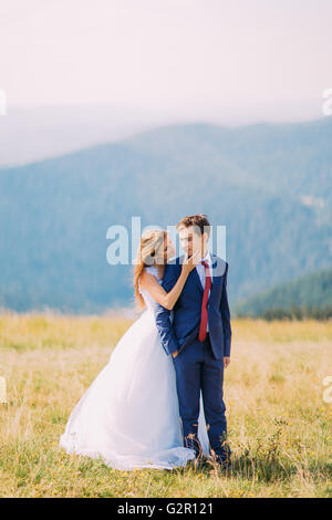 I giovani sposi in posa sul soleggiato Campo con lontane colline di foresta come sfondo. Bella sposa è a baciare i suoi pregi sposo Foto Stock