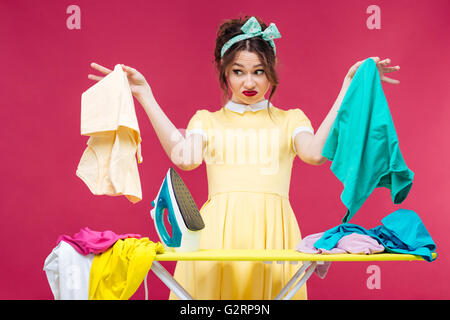 Irritato infelice giovane donna holding e la stiratura di panni su sfondo rosa Foto Stock