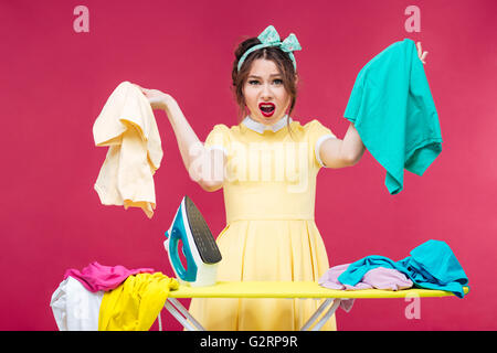 Deluso arrabbiato giovane donna abiti di contenimento e urlando vicino asse da stiro su sfondo rosa Foto Stock