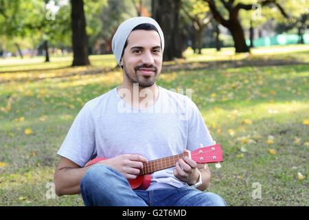 Ritratto di giovane uomo bello suonare il ukelele in un parco. All'esterno. Foto Stock