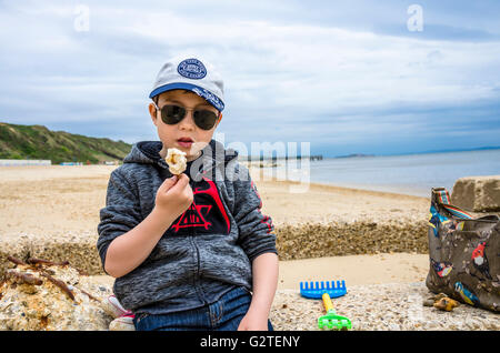 Un ragazzo si siede su una parte di un vecchio per la difesa del mare sulla spiaggia di Boscombe, Bournemouth mangiare un gelato. Foto Stock