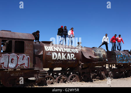 Gli studenti in piedi sulla parte superiore del vecchio motore a vapore con Welcome Dakar graffiti dipinto su di esso, Uyuni treno cimitero, Bolivia Foto Stock