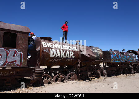 Studente permanente sulla sommità del vecchio motore a vapore con Welcome Dakar graffiti dipinto su di esso, Uyuni treno cimitero, Bolivia Foto Stock