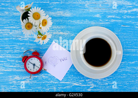 Fiori a margherita, sveglia e la tazza di caffè con ti amo nota su rustiche in legno di colore blu sullo sfondo Foto Stock