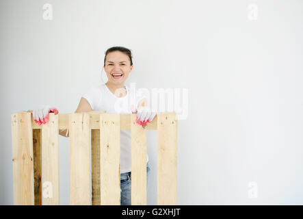 La bella ragazza tiene per mano il pallet in legno. Lei si guarda in una camera e sorrisi. Le sue mani in guanti da lavoro. Foto Stock