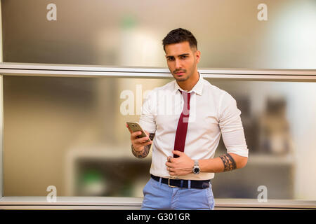 Bel giovane imprenditore nel suo ufficio e di digitazione tramite telefono cellulare, guardando la telecamera, gravi Foto Stock
