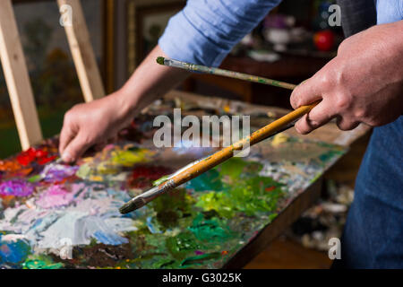 Pittore professionista di contenimento dei pennelli e miscelazione di vernice da paletteknife sulla tavolozza colorata di miscele di olio vernici in una galleria. Foto Stock
