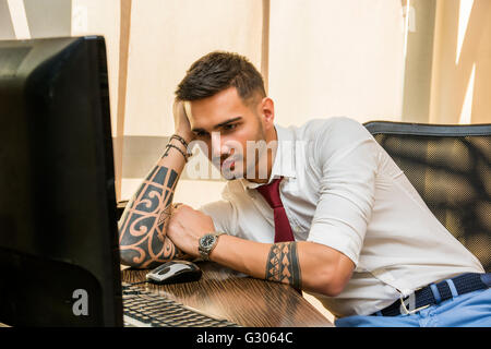 Stanco annoiato giovane imprenditore seduto alla sua scrivania davanti al suo computer sbadigli, tenendo la sua testa con la sua mano e gli occhi clos Foto Stock