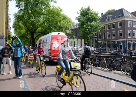 I ciclisti in Bike City Amsterdam corsa lungo il canal road sul luminoso giallo noleggiato biciclette. Foto Stock