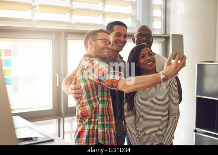 Il gruppo di quattro diversi allegro co-lavoratori tenendo autoritratto al piccolo ufficio di fronte a finestre luminose Foto Stock