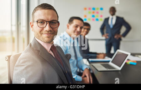 Bel uomo barbuto indossare tuta e cravatta con tre professional co-lavoratori in riunione a un tavolo per conferenza di fronte a un grande Foto Stock