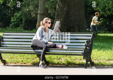 Una giovane donna che legge una rivista su una panchina di parco a Hyde Park, City of Westminster, Londra, Inghilterra, Regno Unito Foto Stock