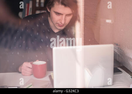 In attesa di ispirazione. Considerato giovane uomo seduto al suo posto di lavoro, bere caffé e la riflessione in vetro Foto Stock