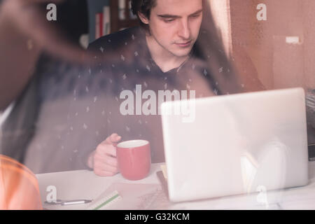 In attesa di ispirazione. Considerato giovane uomo seduto al suo posto di lavoro, bere caffé e la riflessione in vetro Foto Stock