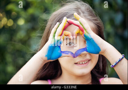 Sorridente bambina con le mani dipinte di realizzare una forma di cuore di fronte il suo occhio Foto Stock