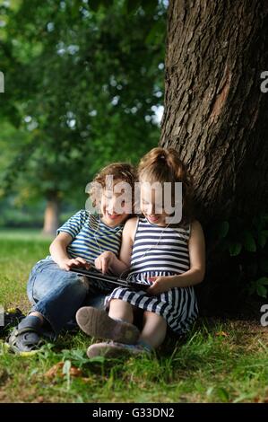 Il ragazzo e ragazza di 7-8 anni look sorridente nel tablet. I bambini seduti dorsi pendente contro un tronco di un grande albero. Essi wi Foto Stock