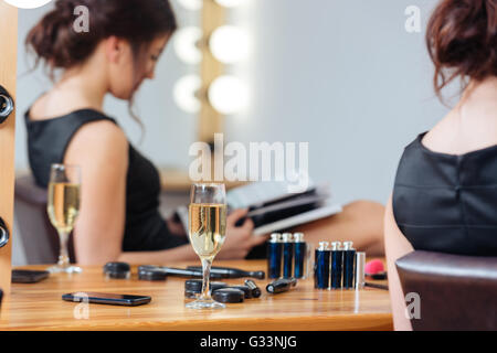 Primo piano della coppa di champagne in piedi sulla tavola vicino al giovane donna rivista di lettura in spogliatoio Foto Stock