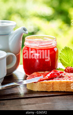 Fetta di pane con il vasetto di marmellata di fragole, tè in tazzina, teiera e fragole fresche e foglie accanto ad esso su un tavolo di legno. Il GRE Foto Stock