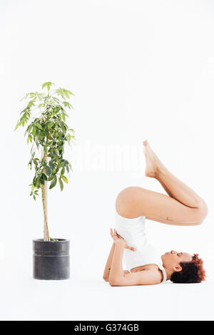 Attraente americano africano giovane donna facendo yoga bilanciamento pongono vicino al piccolo albero in pentola su sfondo bianco Foto Stock