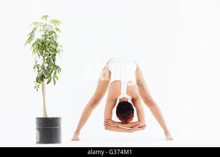 Flessibile americano africano giovane donna stretching e esercizio di yoga vicino al piccolo albero in pentola su sfondo bianco Foto Stock