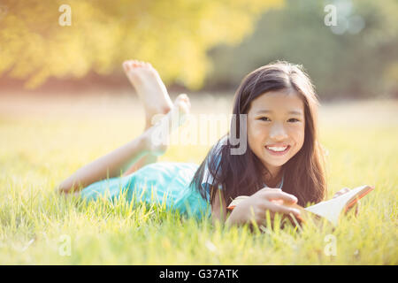 Ritratto di sorridente ragazza distesa su erba e libro di lettura in posizione di parcheggio Foto Stock